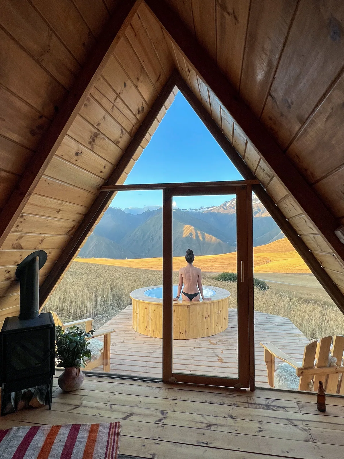 Dall'interno di una baita di legno, la vetrata incornicia Samira nella vasca calda davanti alla valle